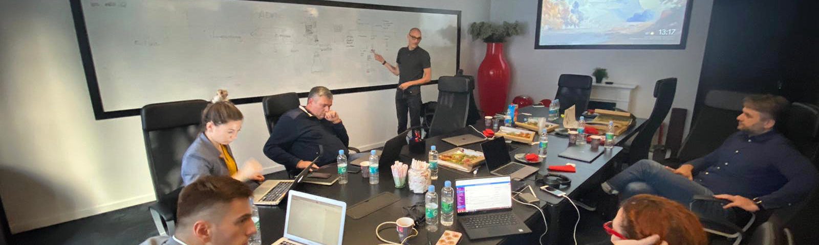 Person pointing to a whiteboard presenting to a room of people sat round a table