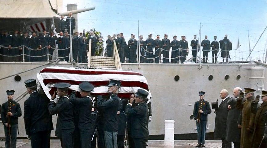 A colorized version of a photo of the Unknown Soldier from World War I as the remains are carried from the USS Olympia on November 9, 1921. To the right of the photo General of the Armies John J. Pershing salutes.