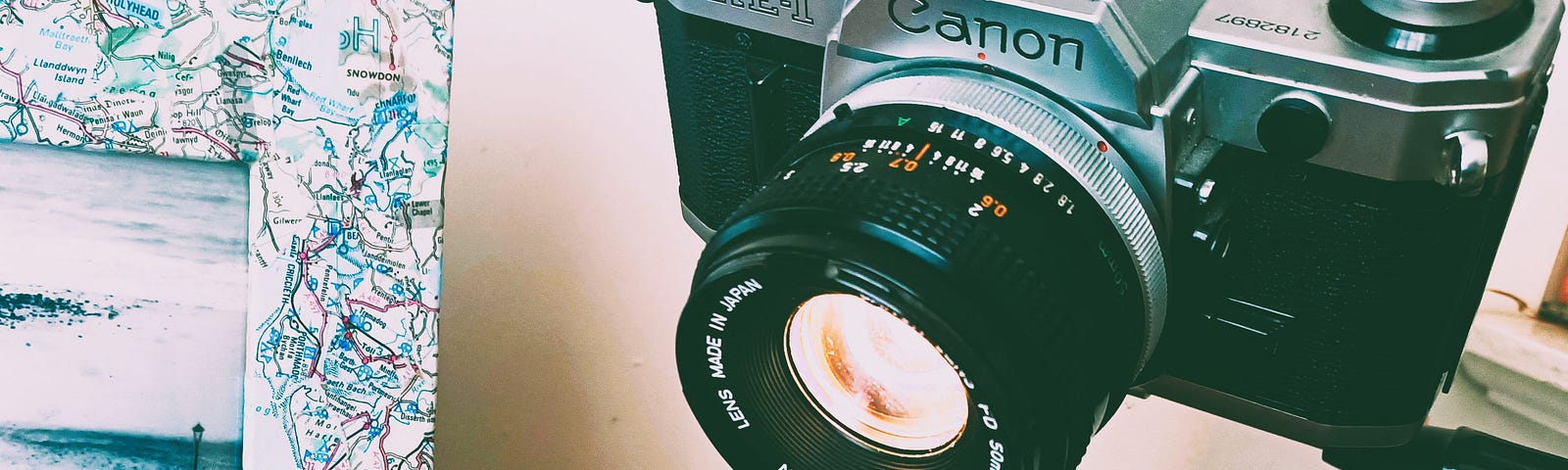 1970s SLR camera on a small tripod on a desktop being used as a desk lamp. There is a picture frame decoupaged in maps to the left with a black and white photograph of the seaside.