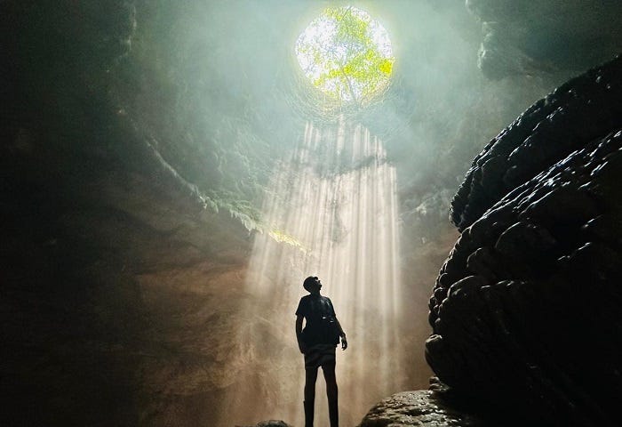man standing in dark cave looking up at sun’s rays streaming down from a hole high overhead