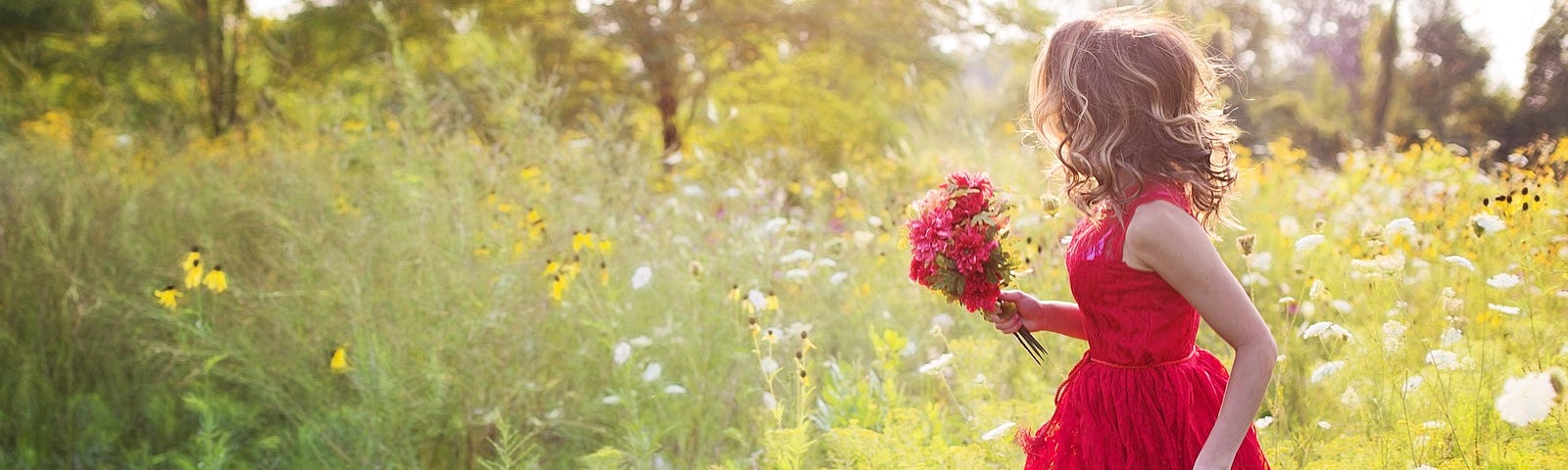 A sunlit field with a young woman in a red dress picking flowers.