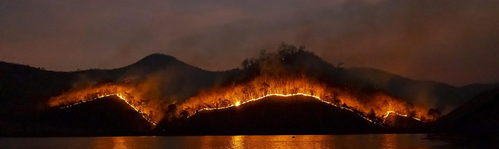 A lake in the foreground reflects the light from a nighttime forest fire on a distant mountain.