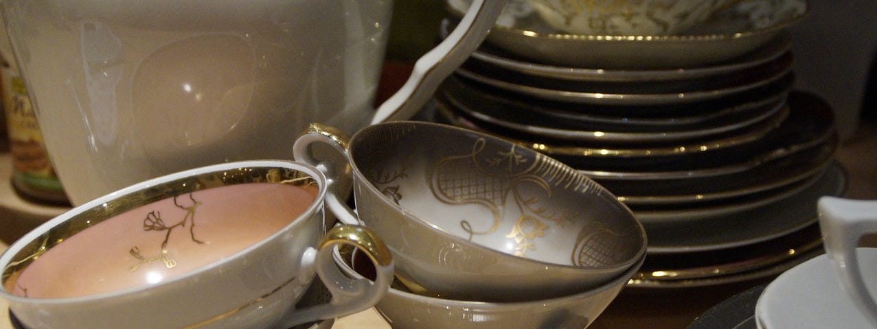 An antique porcelain tea service set, white with gold scrolls. Set includes four teacups in front, a stack of plates in the back, and a pitcher.