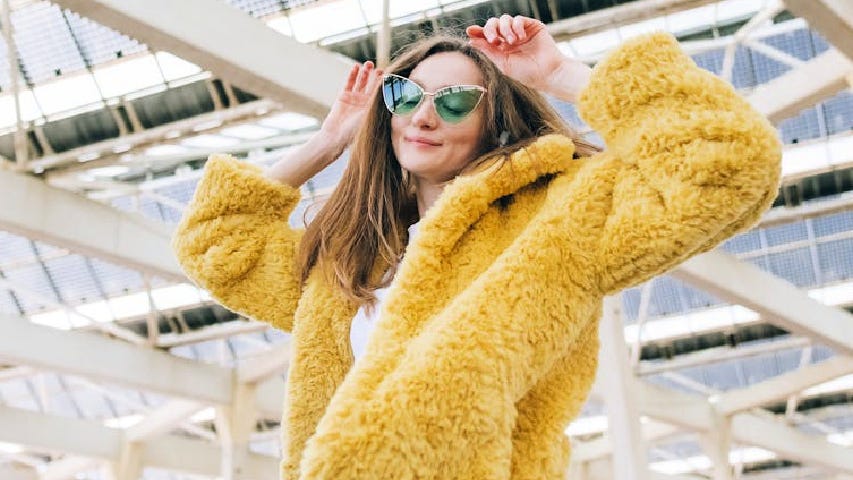 A woman wearing a yellow fake fur coat, green lensed sunglasses, and feeling herself.