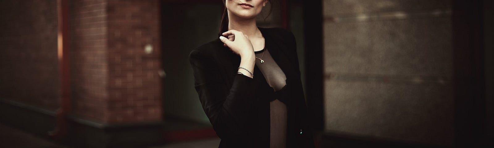 A business woman stands in a brick walled hallway wearing black blazer and sheer blouse.