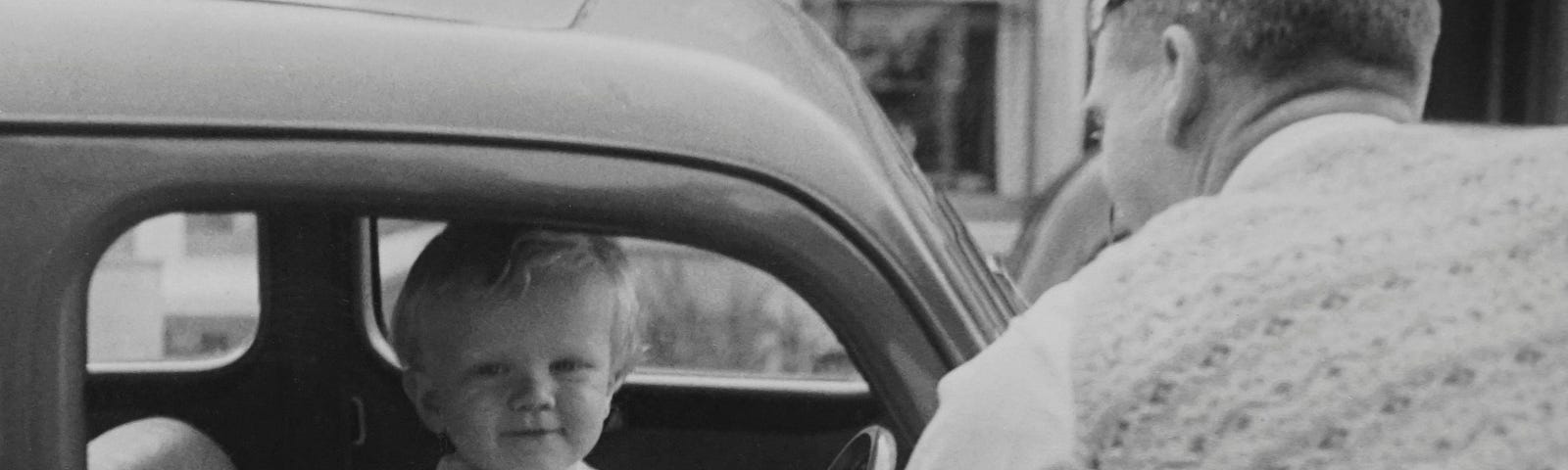 Man looking into an old car with a small boy at the driving seat looking at the camera smiling. Black and white photo.