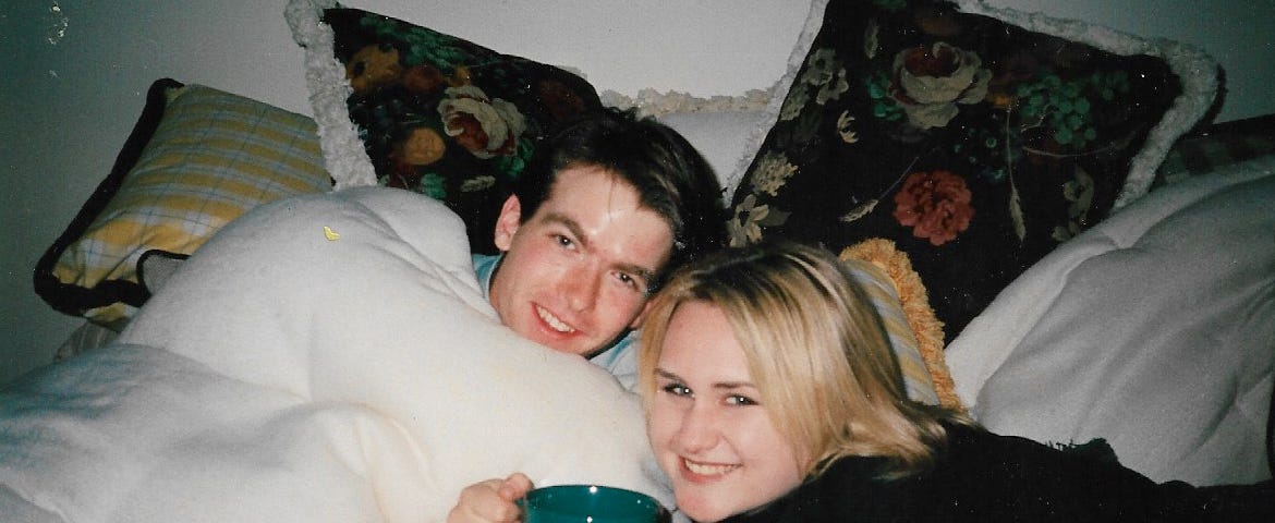 Image of two teenagers fully dressed under the covers of a bed. One is holding a large coffee cup.