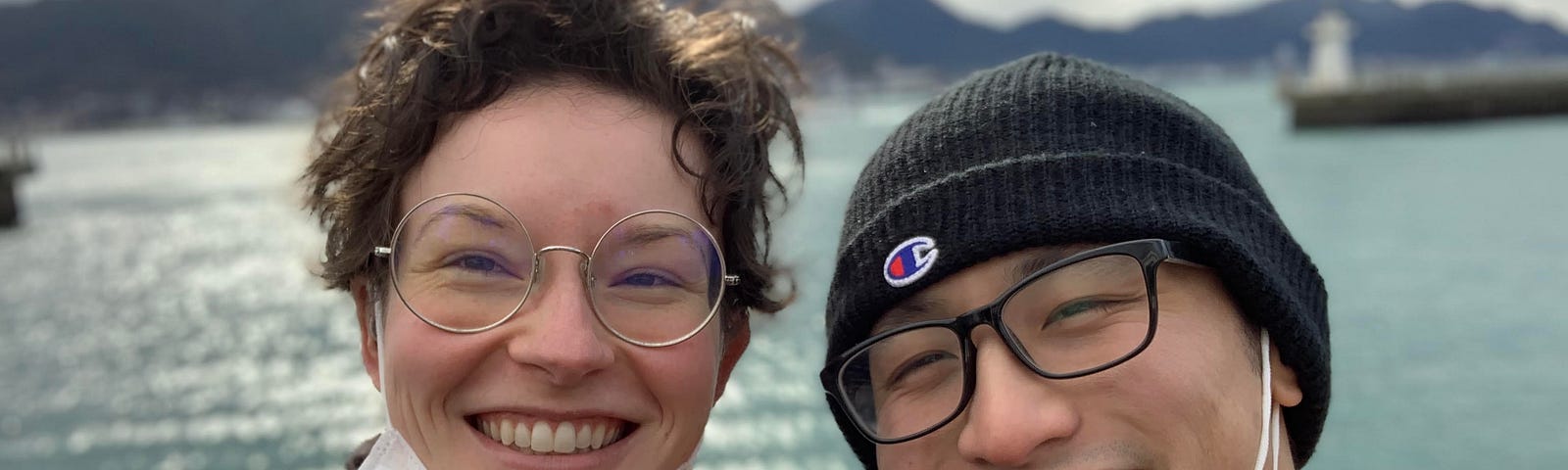 A selfie of my husband and I smiling in front of Kamon Wharf, a little cove in Shimonoseki, Yamaguchi prefecture. I am on the left: a white woman with brown curly hair in an overgrown short cut. I am wearing round silver frame glasses, a mask lowered to my chin, and am bundled up in a black scarf and coat. My husband is on the left: an Asian man wearing a black beanie, a lowered mask, and a bright yellow zip-up coat.