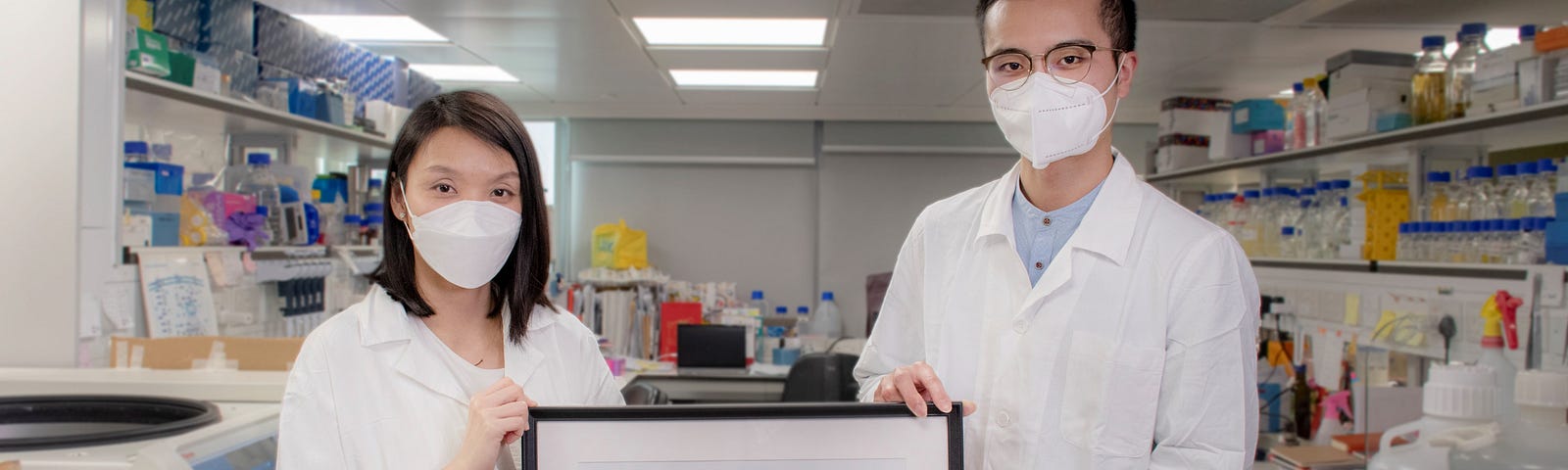 Dr Wong and Vincent stand in a laboratory holding a framed sketch of their colleagues