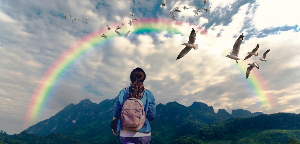 A woman with a backpack walking along the path to mountain and looking rainbow and doves in the sky.
