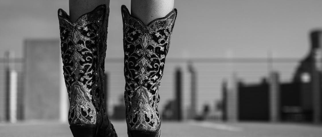 A grayscale image depicts a pair of dark cowboy boots on a person’s feet, trimmed with elaborate silver filagree designs, as they walk towards a blurred cityscape.