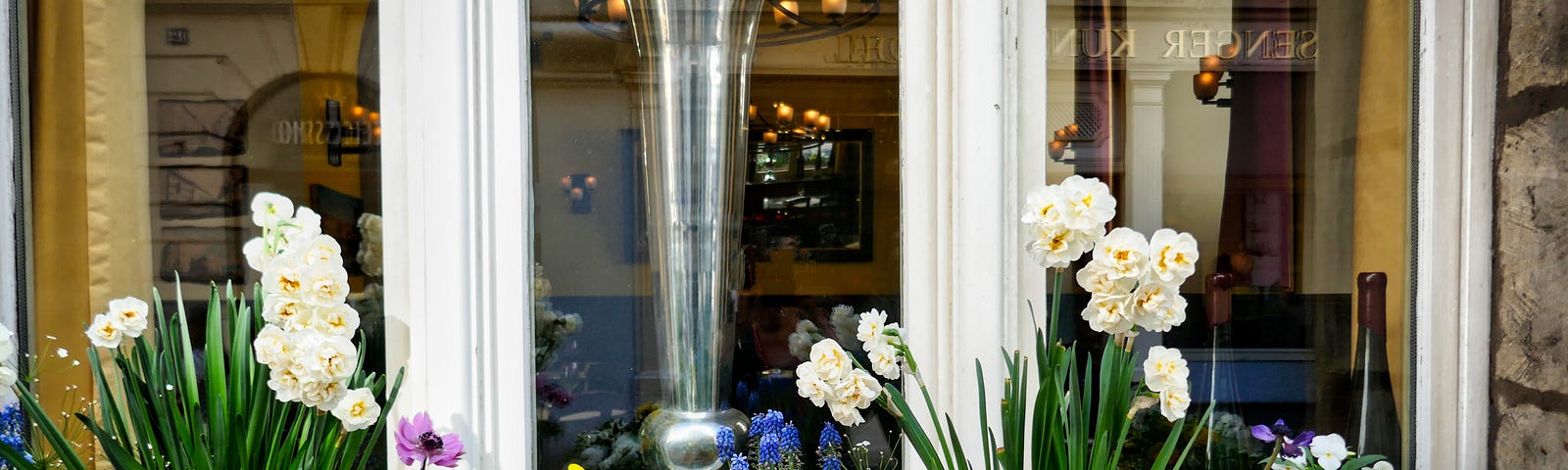 A window in front of a small shop in Germany with a window box full of blooming flowers. You can see a tall glass vase in the window and furnishings in the room.