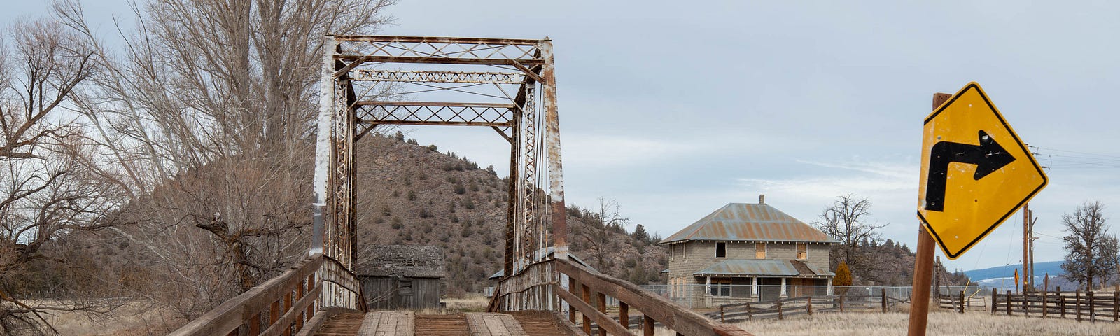 An old one-lane wood-and-iron bridge across a small river on a rural two-lane dirt road, with a farmhouse in the background.