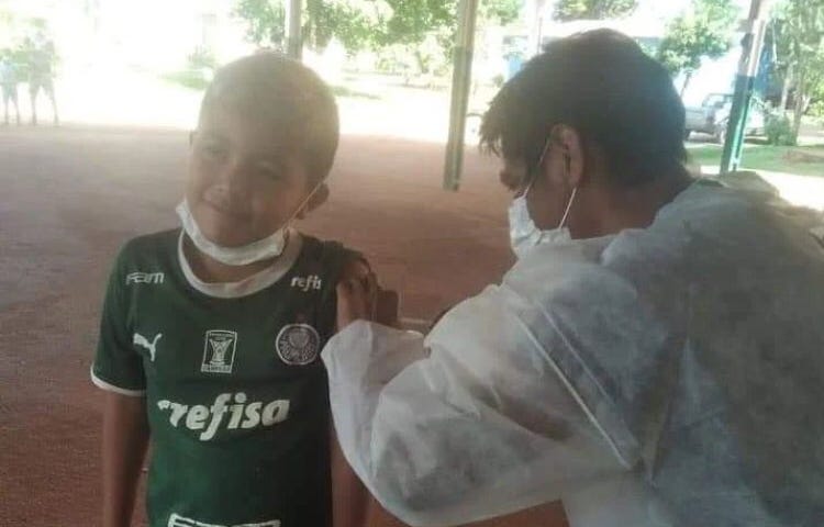 Criança indígena recebe vacina contra a covid-19 vestindo camisa do Palmeiras. Foto mostra ação realizada num lugar com piso de terra batida. Enfermeiro veste jaleco branco, máscara e óculos.