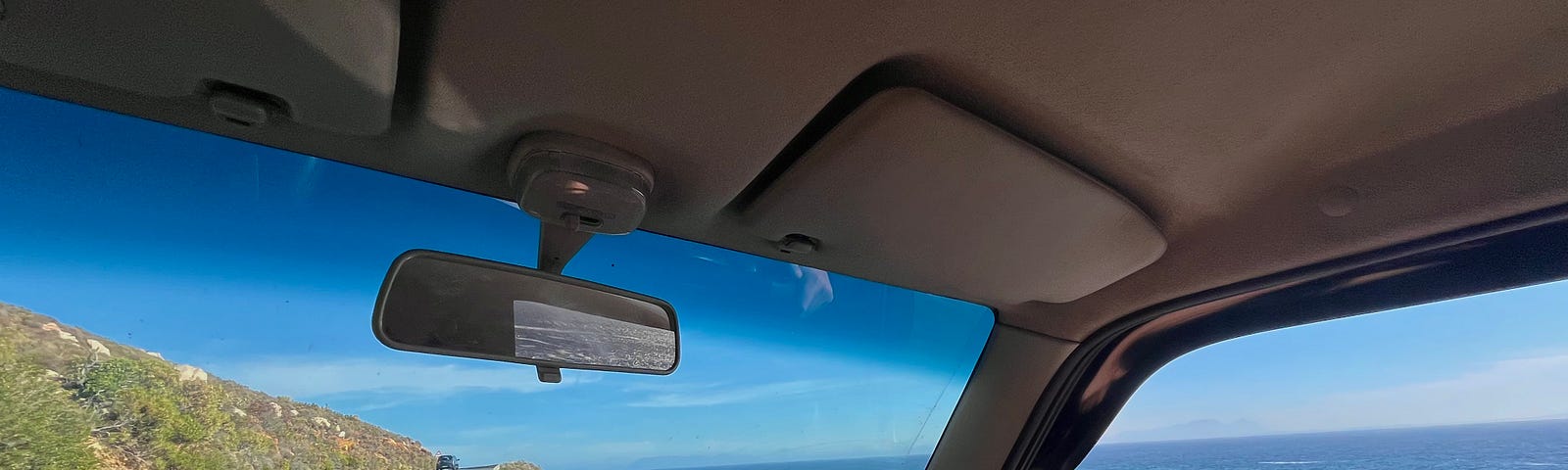 Image out of a car containing the road going forward and the ocean to the left. A hand is on the steering wheel.