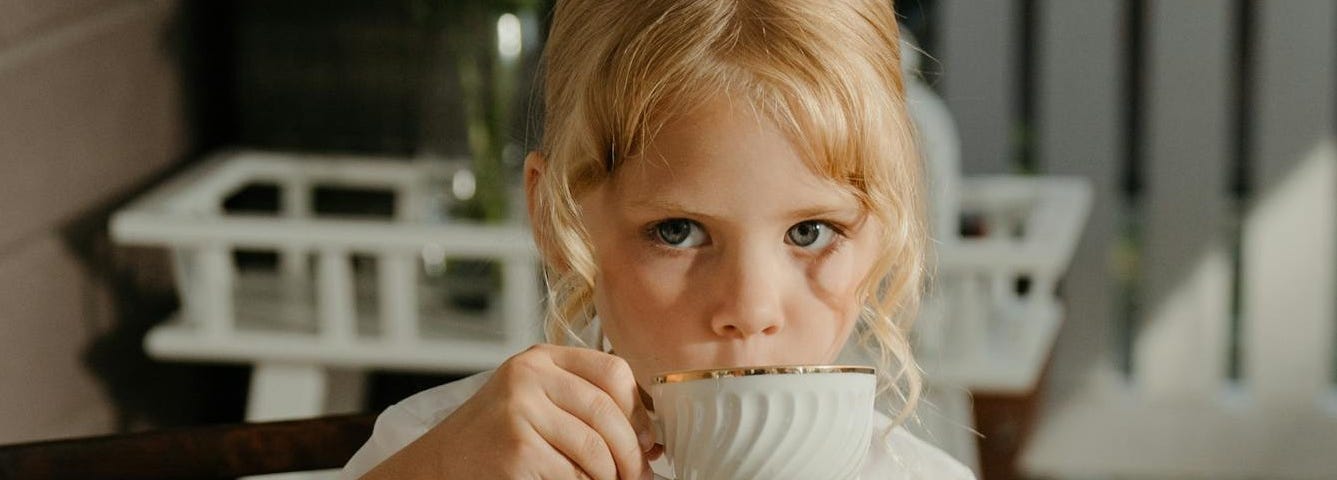 A young girl with blonde hair sits at a table, holding a white teacup with gold trim in one hand and a slice of layered cake in the other. She wears a white blouse with intricate detailing. The background shows a cozy, partially shaded patio area with wooden furniture and soft lighting.