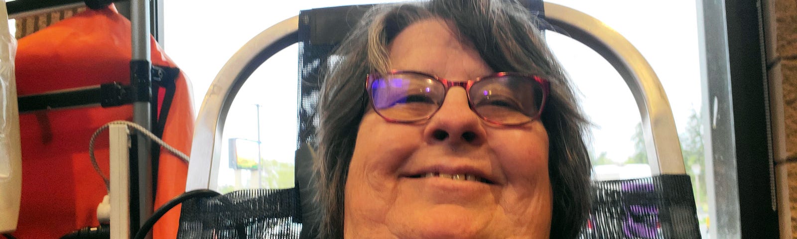 A smiling person with purple glasses is taking a selfie while reclining in a chair, possibly during a blood donation procedure. Behind them, medical equipment can be seen, which suggests a healthcare setting.