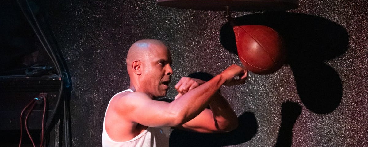 A man with brown skin and a shaved head wears a white tank top. He punches a red speed bag, practicing boxing.