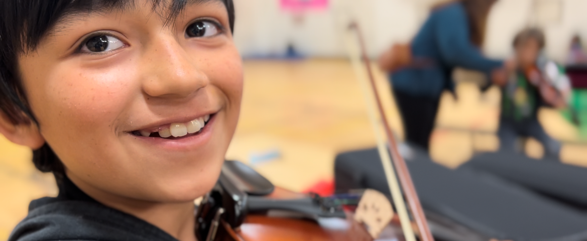 A child playing a violin smiles at the camera.