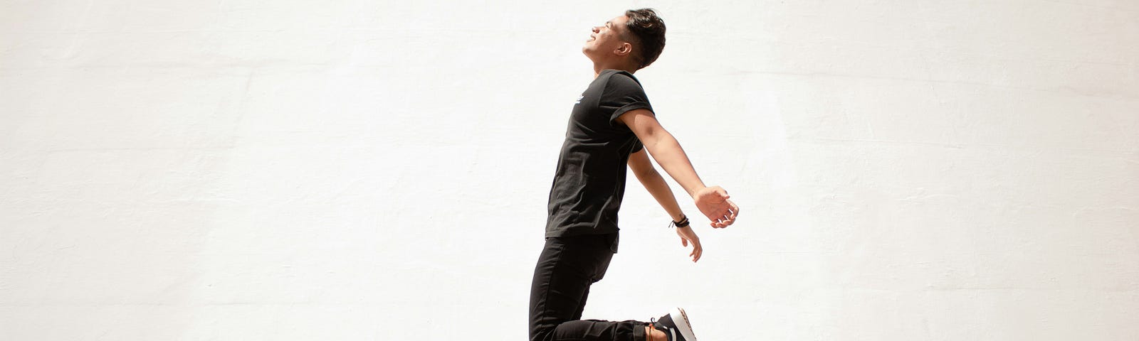 A man dressed in all black, jacket and pants, leaps against a stark white concrete wall in the bright light of day