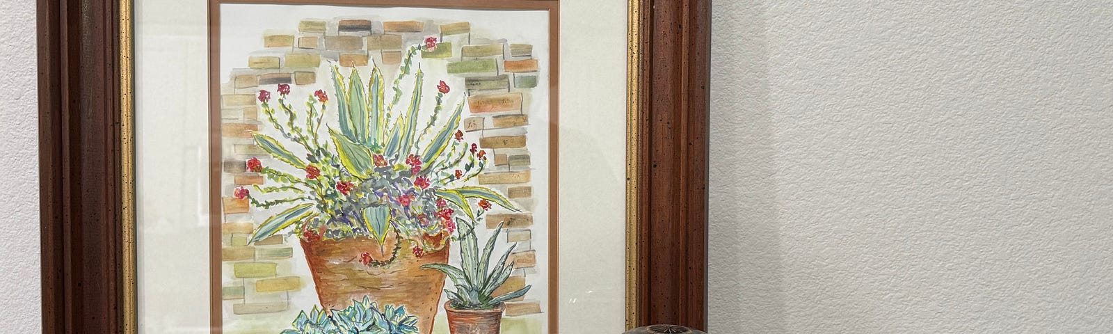 A large framed watercolor of 3 succulents and a black and gold urn sit on a gray side table