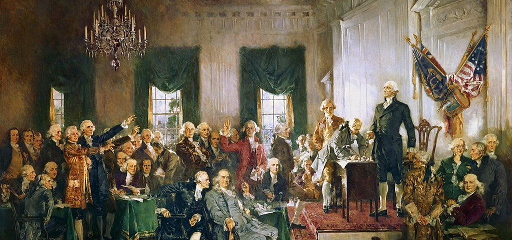 A painting depicts an ornate room full of men. George Washington stands on a platform at right next to a man who signs the Constitution that is laid on a desk. Four flags are displayed on the wall at right.