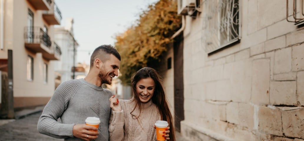 A laughing couple walk closely together down an old European street holding coffee.