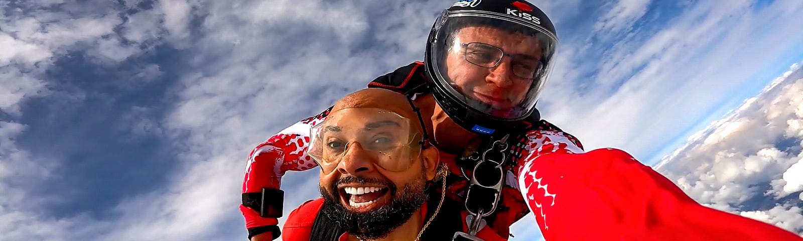 Two skydivers in tandem, the foreground skydiver grinning widely, as they freefall against a backdrop of bright blue sky and soft white clouds, embodying the thrill of skydiving.