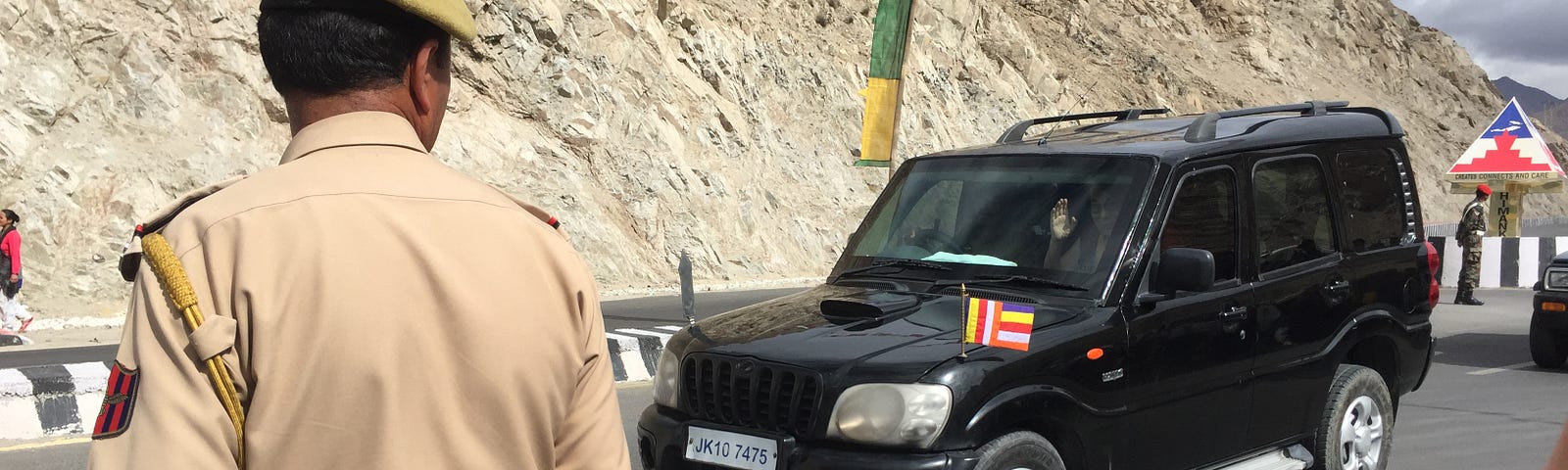 The Dalai Lama is waving his right hand in black SUV. The SUV has a small prayer flag with multi color stripes in front. There is a policeman standing in front to manage the crowds.