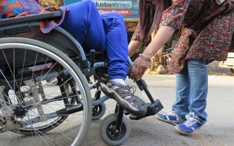 Picture of a person in a wheelchair, and another person standing, who appears to be assisting the first. Their faces are not in the frame. Courtesy of the author.