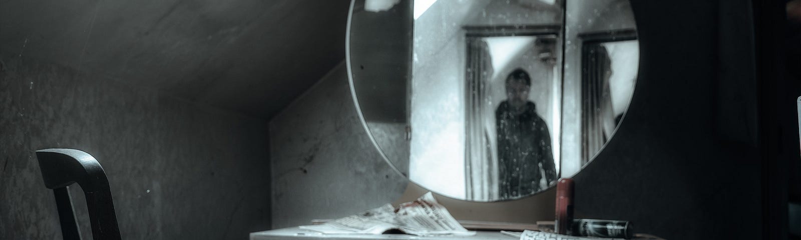 Gloomy dark picture with little colour of a chair in front of an old dressing table with a round mirror on top. The room appears to be in an attic and is dusty. In the mirror a man is seen entering from a doorway, giving the picture an eerie feeling.
