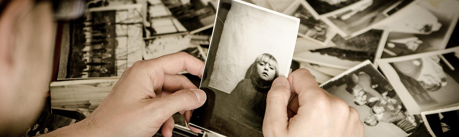 In a black an white photo male hands hold a photo of a young boy amidst a pile of other photos.