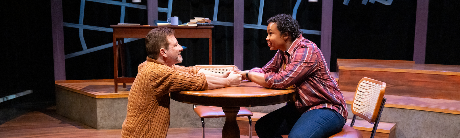 In a photograph from THE WANDERERS, a man in a cardigan kneels at a round table and smiles, holding the hands of a woman in a plaid shirt sitting at the table.