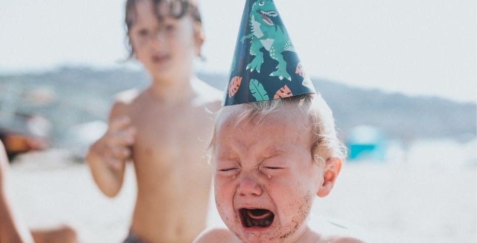 A baby with a birthday hat is crying at the beach