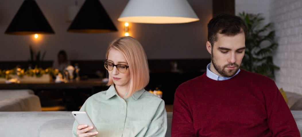 A couple sit next to one another but are distracted by their phones. “Your Partner Doesn’t Know How to Communicate”