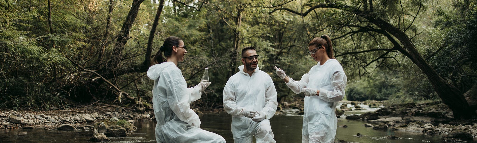 Three people in protective suits take water samples from a shallow river in a green forest.