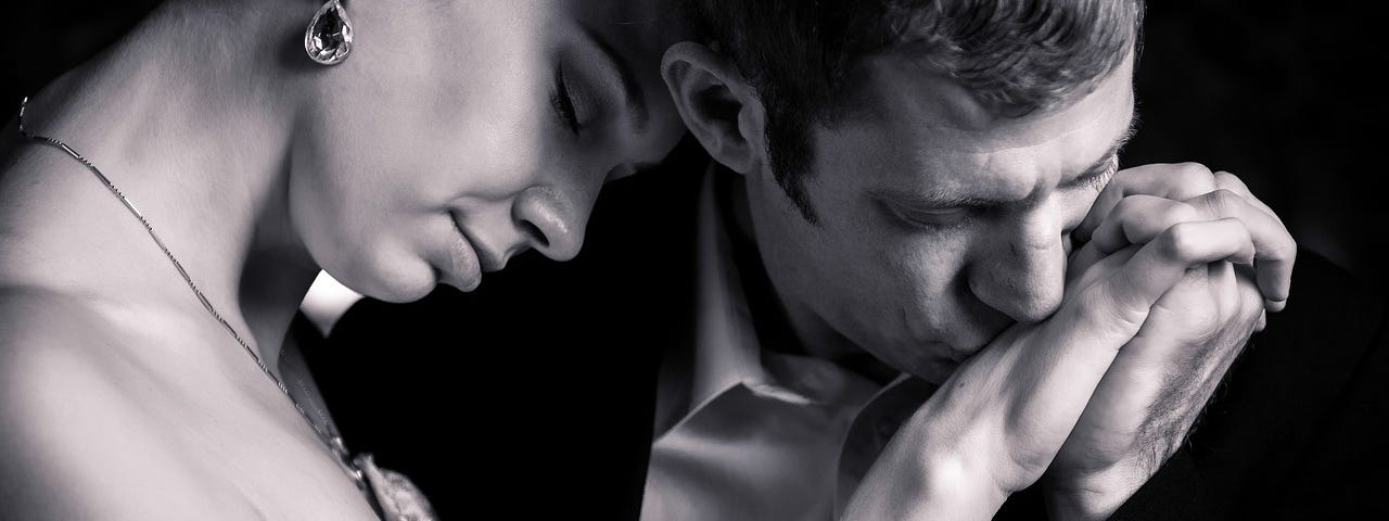 A black and white photo of two lovers, as he places a tender kiss on the back of her hand.