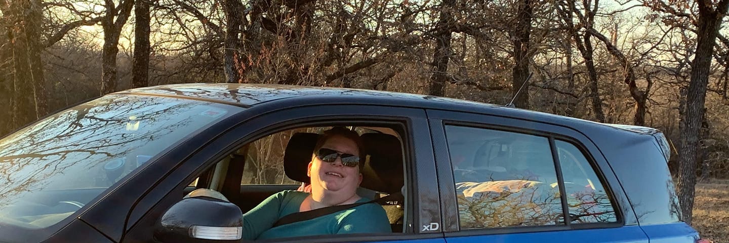 An obese white woman with short brown hair and a teal shirt sits behind the wheel of a blue and black hatchback vehicle.