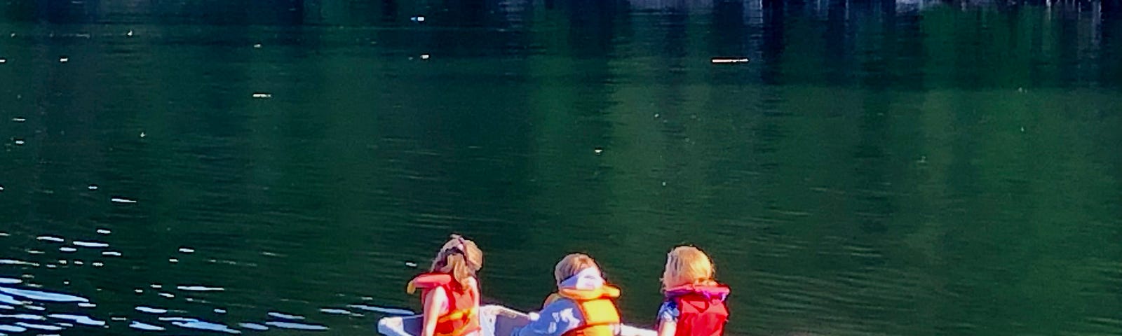 A small green rowboat holds three children one of whom is rowing. Trees and a rocky shoreline are in the background