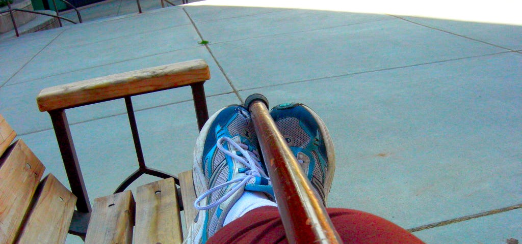 A wooden stick lying on top of a pair of legs in brown pants and feet in blue and pink sneakers lengthwise along a wooden bench.