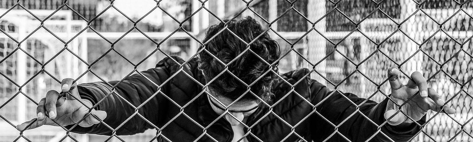 A black & white photo of someone behind a chain link fence, head bowed and hands gripping the fence.