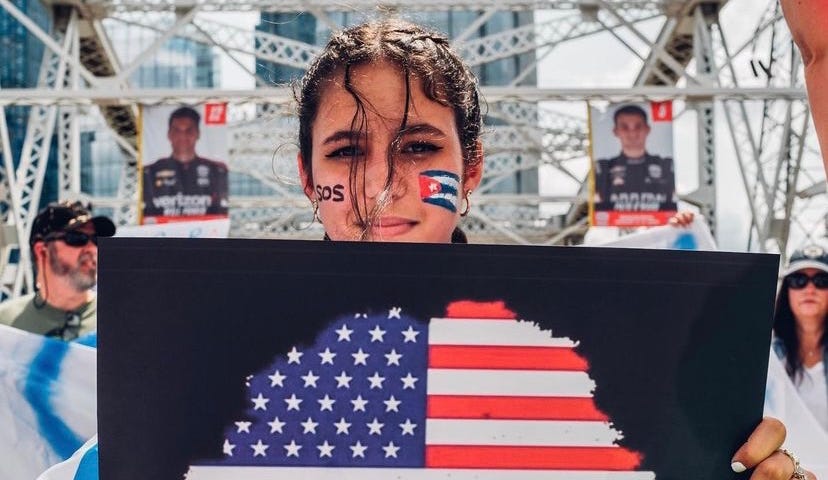 Image by @tony_sarria on Instagram of a woman at an #SOSCUBA protest in Nashville, TN holding a poster of a tree facing north imprinted with the American flag and the words “American Grown with Cuban Roots.”