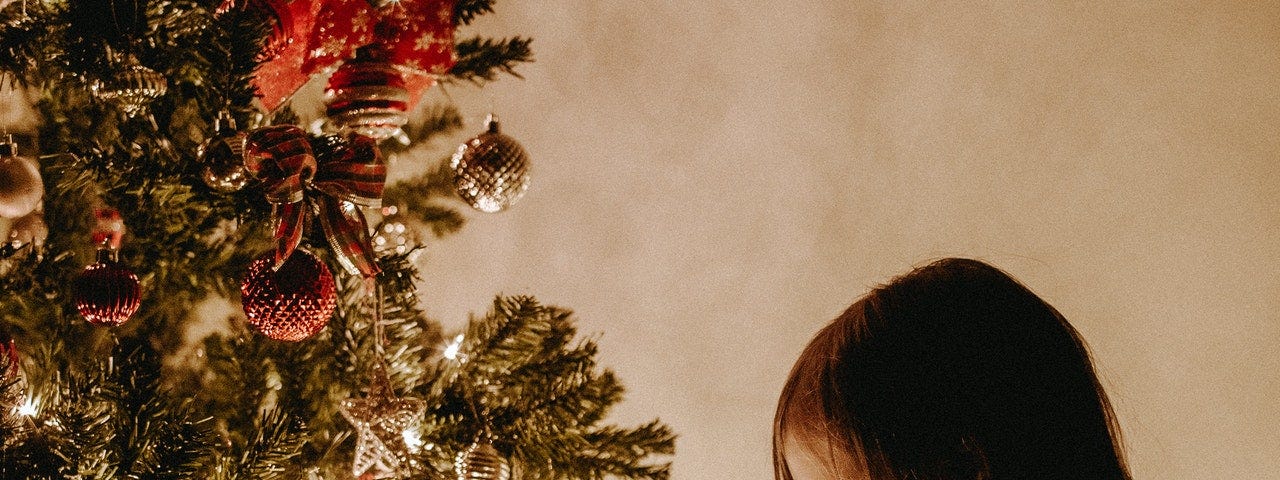 A young girl sits beside a glowing Christmas tree. She is writing a note on a piece of paper.