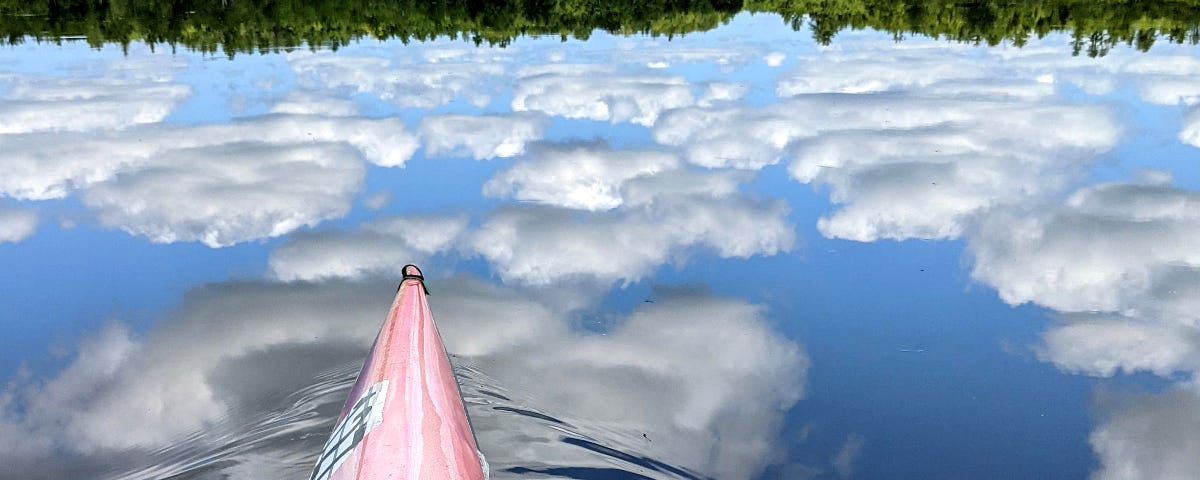 The bow of a kayak creates tiny ripples in an otherwise placid pond that vividly reflects the puffy white clouds in the sky above.