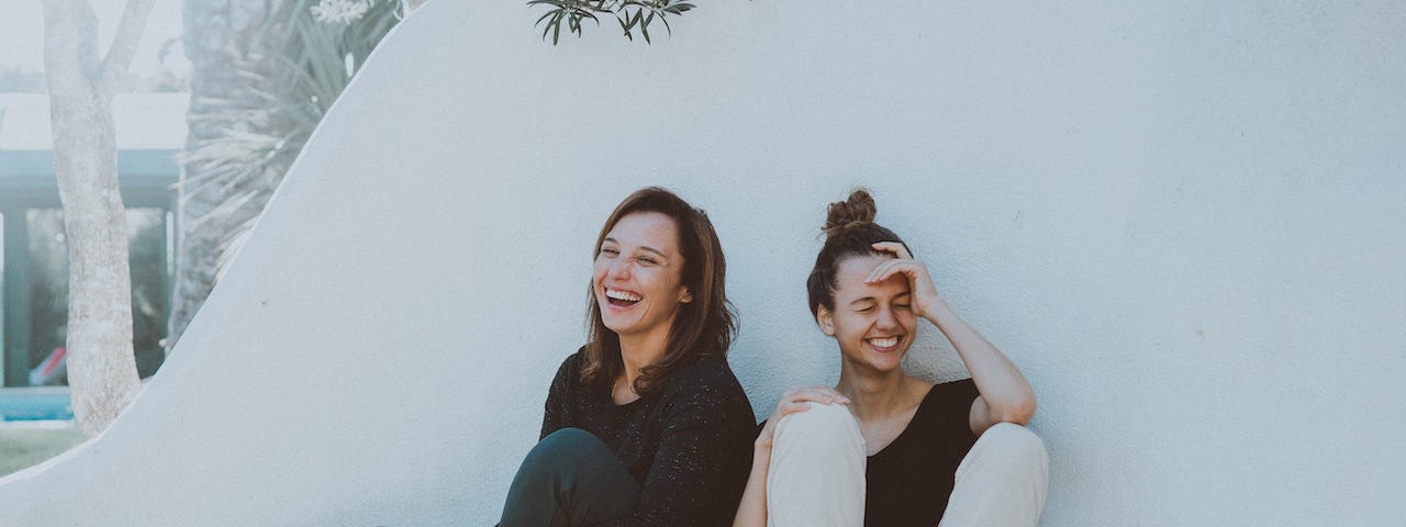 Two female travelers sitting against a white wall laughing.