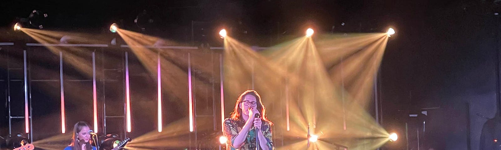 A man with long brown hair stands on stage and sings into a microphone. He wears a patterned long-sleeved top and jeans. Behind him is a guitar player in a blue dress and a keyboard. Golden lights illuminate the man from behind.