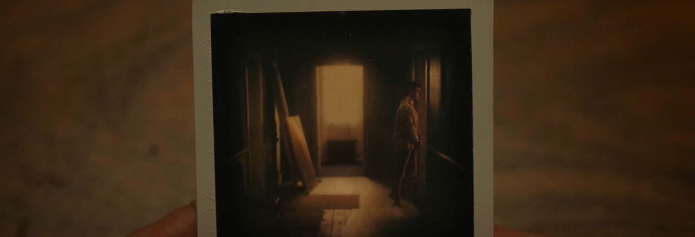 A close-up of a Polaroid photograph held in Philip’s hands, showing a young Philip standing alone in a dim hallway, bathed in warm light coming from an open door at the far end.