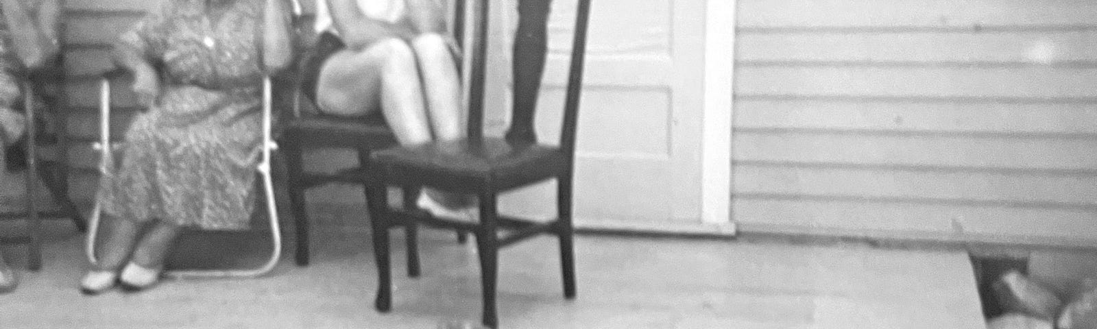 Concrete porch with chairs along a wall of a house. An elderly woman is in the middle and a younger woman is sitting on a chair at the end. A fat baby boy is sitting on the lower step.