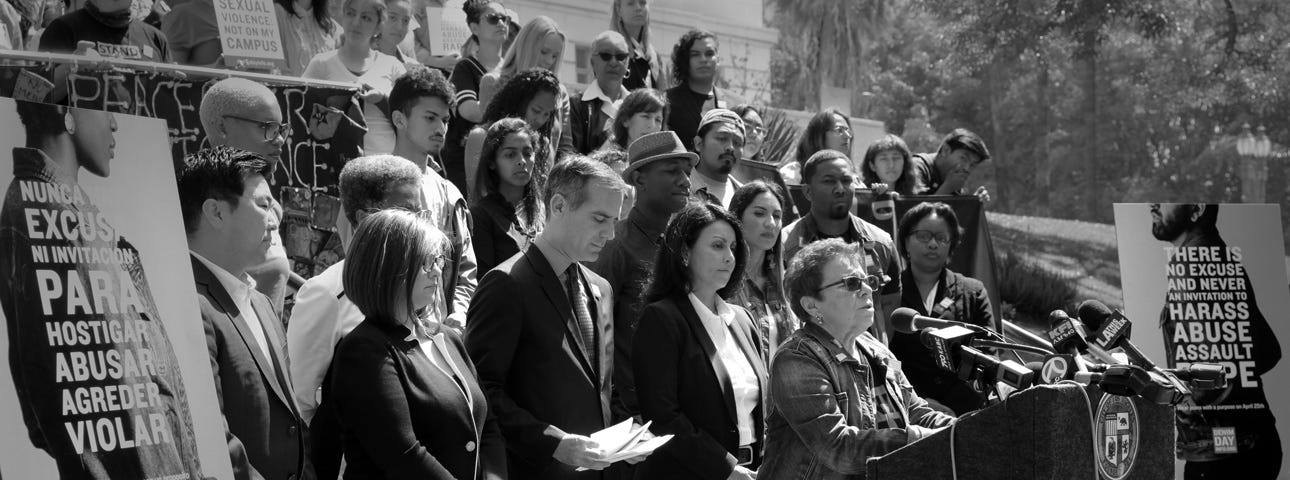 A woman in all denim stands speaking behind a podium. Behind her, a large crowd stands in solidarity, many of which are also wearing denim.