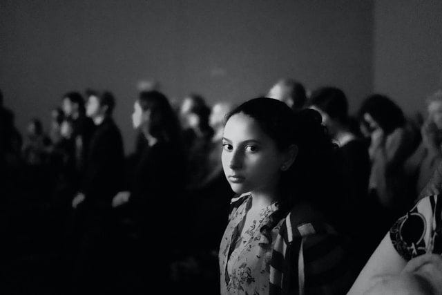 Black and white photo with a young girl in focus glancing toward the camera. Her face is absent of emotion. No smile, no frown either. The room is crowded with people. She looks alone and maybe afraid.